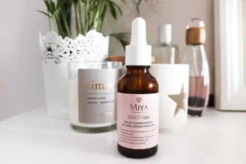 Brązowa buteleczka serum Miya Beauty.lab z pipetką na jasnoróżowej etykiecie, na tle świecy i roślin.