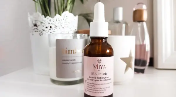 Brązowa buteleczka serum Miya Beauty.lab z pipetką na jasnoróżowej etykiecie, na tle świecy i roślin.